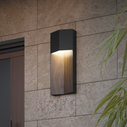 The Artika Howel modern LED indoor-outdoor wall light 5 CCT, featuring dark and wood-like finishes, emits warm light and is mounted on a beige stone wall near a wooden door and green plant.