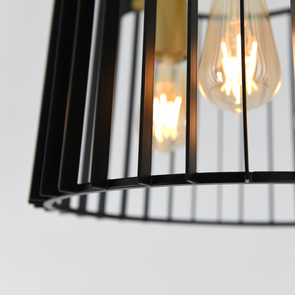 Close-up of a modern black metal light fixture with exposed filament bulbs glowing warmly, viewed through vertical metal bars against a plain, light background.