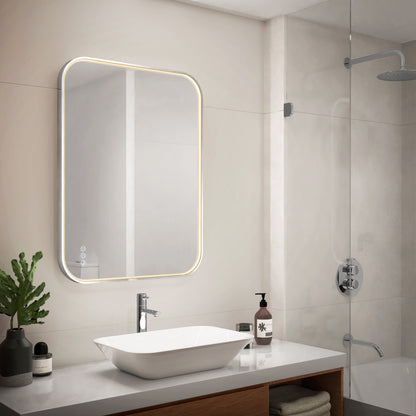 Modern bathroom with a rectangular backlit mirror above a white sink, silver faucet, soap dispenser, and potted plant on the countertop. A glass shower enclosure with a rain showerhead is visible on the right.