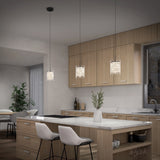 A modern kitchen with light wood cabinets, a white island with two cushioned bar stools, pendant lights hanging above, a vase with greenery on the countertop, and a window above the sink.
