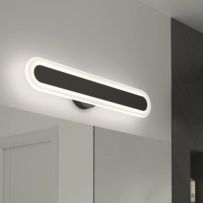 The Artika Pista modern LED bathroom vanity light 5 CCT, featuring an oval black design with a glowing white border, is mounted above a bathroom mirror on a white wall beside dark tiles.