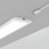 A rectangular LED ceiling light glows brightly on a gray ceiling. Above it, a white USB-C cable with a connector is positioned, appearing ready to plug into the light.