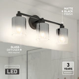 A modern bathroom wall light fixture with three cylindrical glass diffusers, each containing a visible bulb. The fixture has a matte black finish and is mounted above a mirror. Text highlights integrated LED and a 3-year warranty.