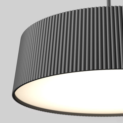 Close-up of the Artika Groove Drum LED contemporary pendant light 3 CCT, a modern black ribbed cylindrical lamp hanging from the ceiling, illuminated and casting a warm glow against a plain grey background.