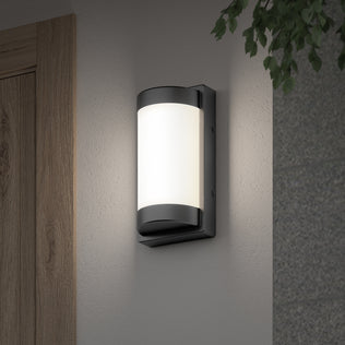 The Artika Fogo modern LED indoor-outdoor wall light 5 CCT is mounted on a light-colored wall near a wooden door, with green leaves in the top right corner.