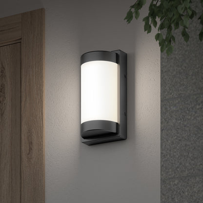 The Artika Fogo modern LED indoor-outdoor wall light 5 CCT is mounted on a light-colored wall near a wooden door, with green leaves in the top right corner.