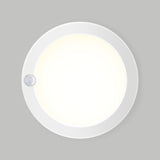 A round, white ceiling light with a small sensor on its edge is mounted on a plain gray wall and emits a bright, even glow.