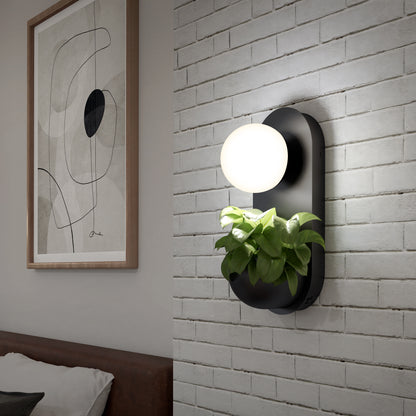 The Artika Bloomfield LED modern mid-century wall sconce 5 CCT, featuring a round white light and a small green plant, is mounted on a white brick wall beside an abstract line art picture above the bed.