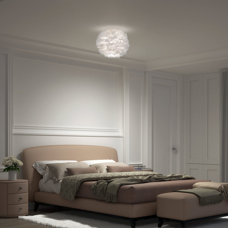 A softly lit bedroom features Artikas Plume LED flush mount light, a beige upholstered bed with green and gray bedding, a matching bench, a nightstand with flowers, and white panel-detailed walls.