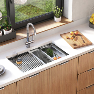 The Artika Newport twin bowl 33 in. stainless steel workstation sink features a pull-down faucet, drying rack with dishes, built-in colander for herbs and fruits, and wooden cutting board with sliced fruit on a white countertop by the window.