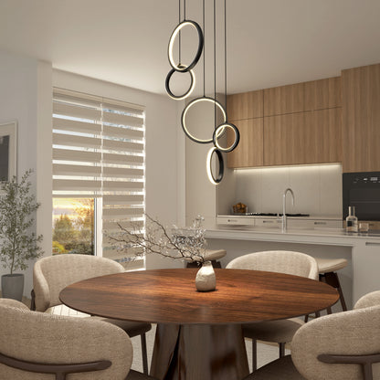 Modern dining area featuring a round wooden table, beige chairs, and the Elmo 5-light LED pendant by Artika. Kitchen with wood cabinets, built-in oven in the background, and sunlight streaming through blinds.