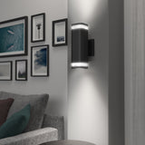 A modern black wall sconce with upper and lower LED lights is mounted on a white wall near a gray sofa. Framed photos and artwork are arranged on the adjacent wall.