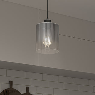 The Artika Haze pendant light with a clear glass shade and visible bulb hangs from the ceiling in a kitchen with gray cabinets and a white subway tile backsplash.