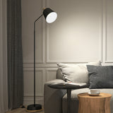 A modern living room with a black floor lamp beside a grey sofa, a small round side table with an open magazine, and a wooden table holding a white cup and saucer, all set against white paneled walls.