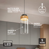 A modern pendant light with a matte black and painted wood finish, smoked glass diffuser, and adjustable height hangs in a kitchen. Icons highlight its integrated LED, energy efficiency, and 5-year warranty.