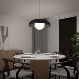 A modern dining room with a round table set for four, black chairs, a black and white pendant light overhead, a plant in the corner, large windows, and minimalist decor on the wall.
