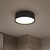 A modern, round black ceiling light with a ribbed texture is installed on a light-colored ceiling in a contemporary room with wooden wall panels and cabinets. The light is turned on, illuminating the area below.