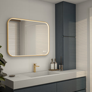Modern bathroom with an Artika Royce Dimmable LED Mirror (3 CCT, horizontal/vertical mount) and touch controls above a white countertop sink. Gold faucet, dark blue cabinets, and decorative bottles stand out in the sunlight.