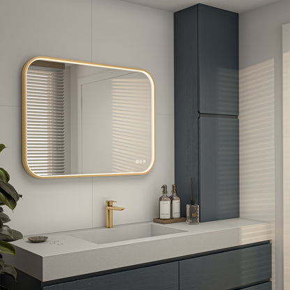 Modern bathroom with an Artika Royce Dimmable LED Mirror (3 CCT, horizontal/vertical mount) and touch controls above a white countertop sink. Gold faucet, dark blue cabinets, and decorative bottles stand out in the sunlight.
