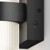 Close-up of a modern wall-mounted light fixture with a ribbed glass shade and a matte black metal base, featuring a round power button and a screw detail visible on the side.