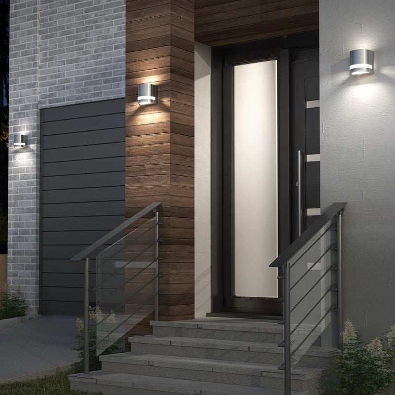 Modern house entrance with concrete steps, metal railings, a tall glass door, and Artika Valor Round LED indoor-outdoor wall lights illuminating brick, wood, and stucco exteriors at dusk.