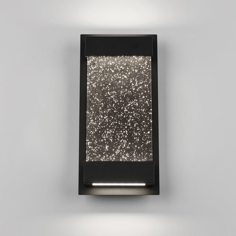 The Artika Essence Glow Box indoor-outdoor LED wall light features a modern rectangular design with a black frame and star-textured illuminated glass, casting soft up-and-down lighting against a light gray wall.