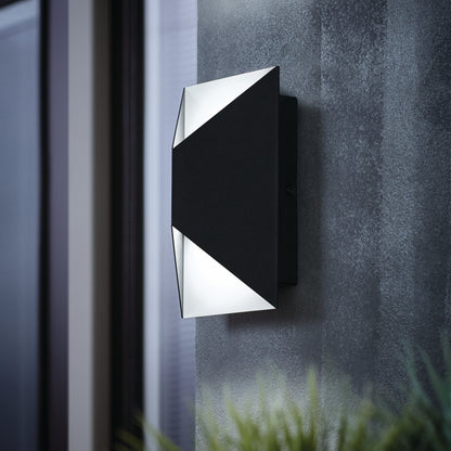 The Artika Tria LED indoor-outdoor wall light, featuring a modern black and white geometric design, is mounted on a textured gray wall and emits soft up-and-down light. A blurred window and greenery appear in the background.