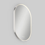 A modern, oval-shaped mirror with a thin metallic frame and built-in LED lighting along the edge, mounted on a plain light gray wall. Three small touch control icons are visible at the bottom of the mirror.
