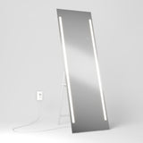 A tall rectangular mirror with two vertical LED light strips stands on the floor, propped up on a white support. A white power cord is plugged into a nearby wall outlet against a plain white background.