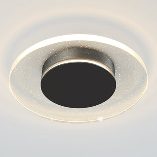 The Artika Essence Disk LED flush mount is a modern ceiling light featuring a black center, transparent outer ring with sparkling specks, and emits a soft white glow.