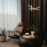 A modern lounge area with a black leather chair, a round ottoman topped with books and potted plants, a tall plant on the side, sheer curtains, and a contemporary ceiling light fixture.