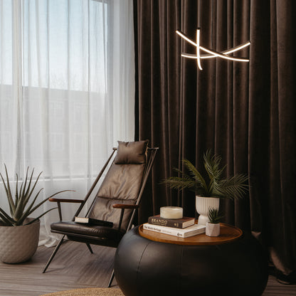 A modern lounge with a leather chair, a round black ottoman topped with books, a candle, and potted plants. Floor-to-ceiling curtains and the Artika Orly modern LED pendant light 5 CCT complete the cozy interior.