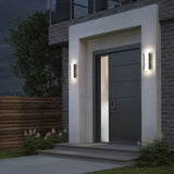 Modern house entrance at dusk with a sleek dark door, illuminated by two rectangular wall lights on either side. Concrete steps lead up to the doorway, and there are green shrubs and a wooden fence nearby.