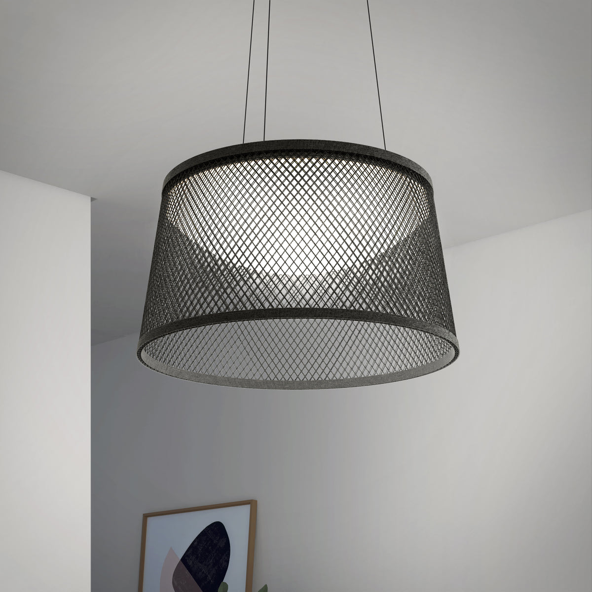 A modern ceiling pendant light with a black mesh drum-shaped shade hangs from two wires in a minimalist room with white walls and a framed abstract artwork visible in the background.