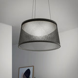 A modern ceiling pendant light with a black mesh drum-shaped shade hangs from two wires in a minimalist room with white walls and a framed abstract artwork visible in the background.