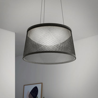 The Artika Adrian LED pendant light 3 CCT, featuring a black mesh drum shade, hangs from the ceiling and casts a soft glow. In the background are white walls, a corner, and part of framed abstract artwork.
