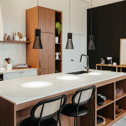 Modern kitchen with a marble island, wooden cabinets, two black chairs, open shelves with plants and kitchenware, and minimal decor. Features Artika’s Kiyo modern LED pendant 3 CCT lights hanging above the countertop.