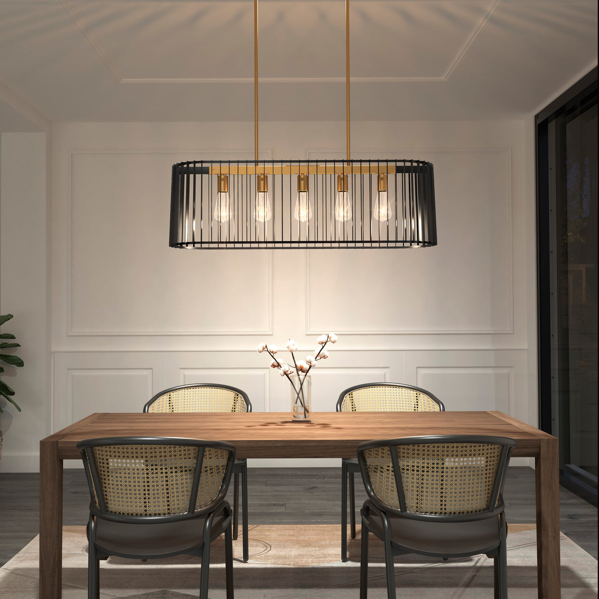 A modern dining room with a wooden table, four rattan-backed chairs, a minimalist glass vase with twigs, and a black and gold rectangular pendant light with exposed bulbs above the table. White paneled walls and a large window.
