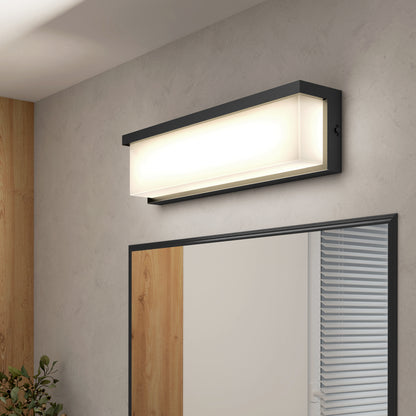 The Artika Bristol modern LED bathroom vanity light 3 CCT, featuring a black frame, casts a soft glow above a large mirror in a modern room with light gray walls, wooden accents, and window blinds.