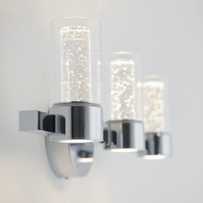 Close-up of the Artika Novello modern LED bathroom vanity light, featuring three bubble-filled cylindrical glass shades on a chrome base, mounted against a white wall.