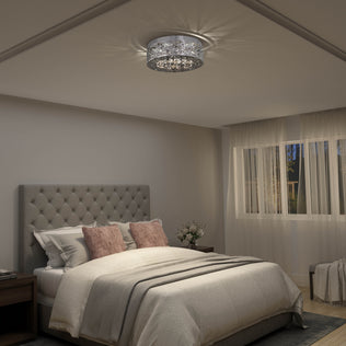 A cozy bedroom features a tufted gray headboard, white bedding, blush pink pillows, and Artika’s Crystal Nest LED flush mount. Sheer curtains cover large windows, and a bedside table holds a small flower vase.