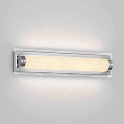 The Artika Glamour LED bathroom vanity light 3 CCT is a modern, wall-mounted fixture featuring a cylindrical textured glass shade and chrome accents, casting a soft, warm glow on a plain light gray wall.