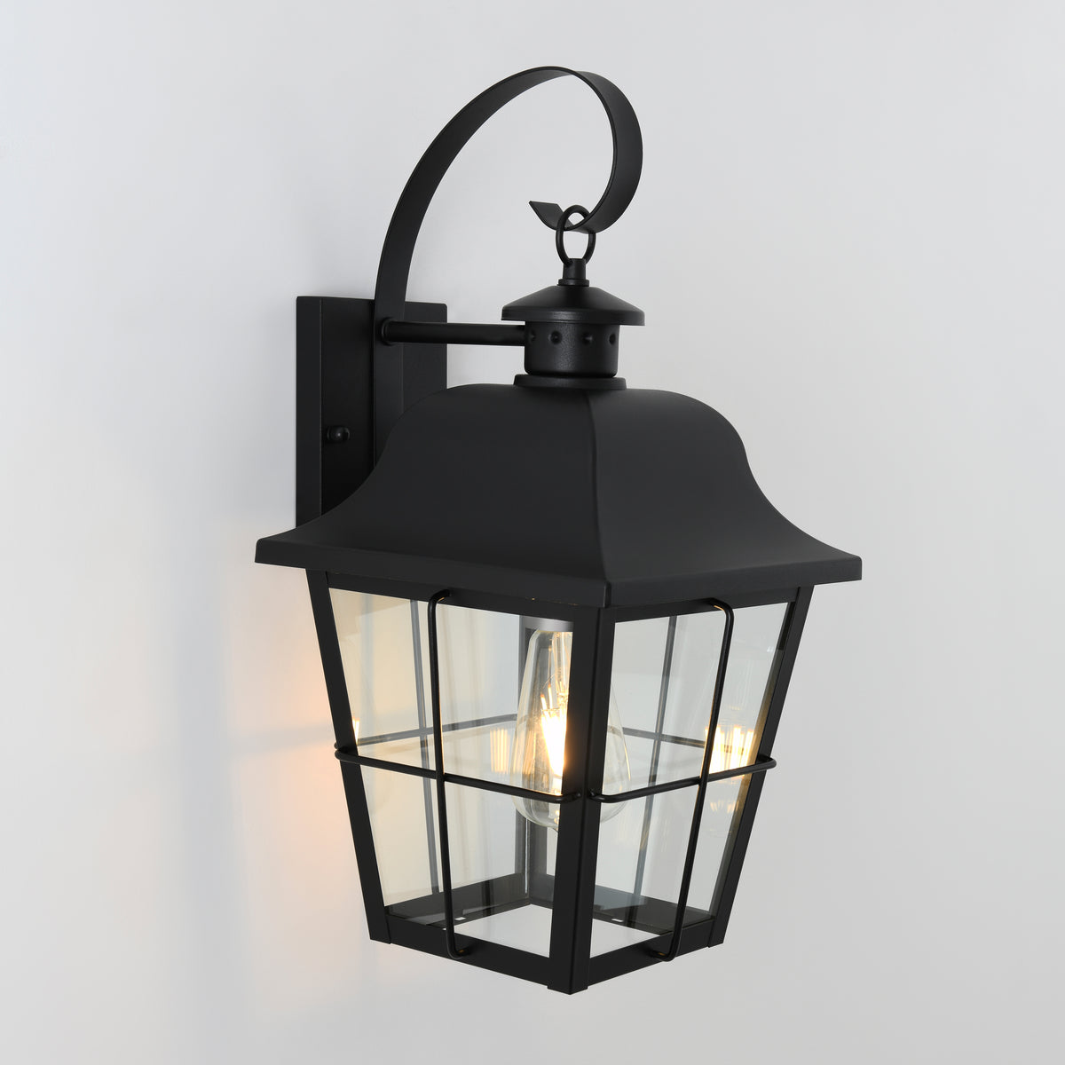 A black metal outdoor wall lantern with clear glass panels, a curved top arm, and a visible glowing bulb inside, mounted on a white wall.