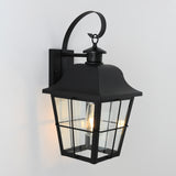 A black metal outdoor wall lantern with clear glass panels, a curved top arm, and a visible glowing bulb inside, mounted on a white wall.