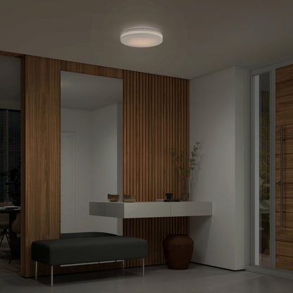 A modern entryway features a wooden accent wall, large mirror, floating white console table, dark bench, ceramic vase, and is softly illuminated by the Artika Hemisphere 15 in. LED flush mount light with 5 CCT.