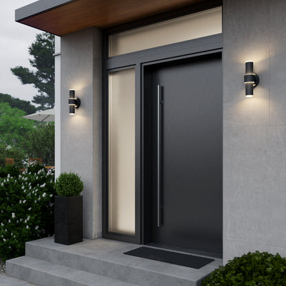 Modern house entrance with a large black door, vertical window, and two Artika Dani modern LED indoor-outdoor wall lights (5 CCT) on either side. Concrete steps lead to the door, with a rectangular shrub planter on the left and nearby bushes.