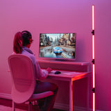 A person wearing headphones sits at a desk playing a racing video game on a monitor. The room is lit with pink and red LED lights, creating a colorful, modern gaming setup.