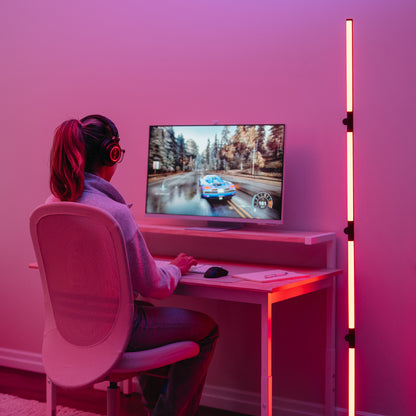 A person wearing headphones plays a racing game at a desk, illuminated by the Artika Fitch smart foldable RGB LED gaming floor lamp in a modern, minimalist room.
