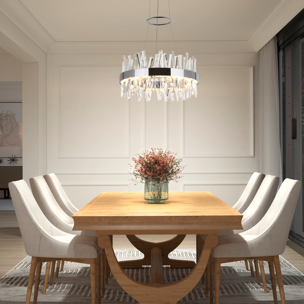 A modern dining room with a wooden table, six upholstered chairs, a vase of red flowers as the centerpiece, a contemporary chandelier overhead, and large windows with sheer curtains.