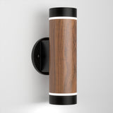 A cylindrical wall sconce with a wood-textured center and black metal ends, mounted on a round black wall plate, emitting light from both the top and bottom.